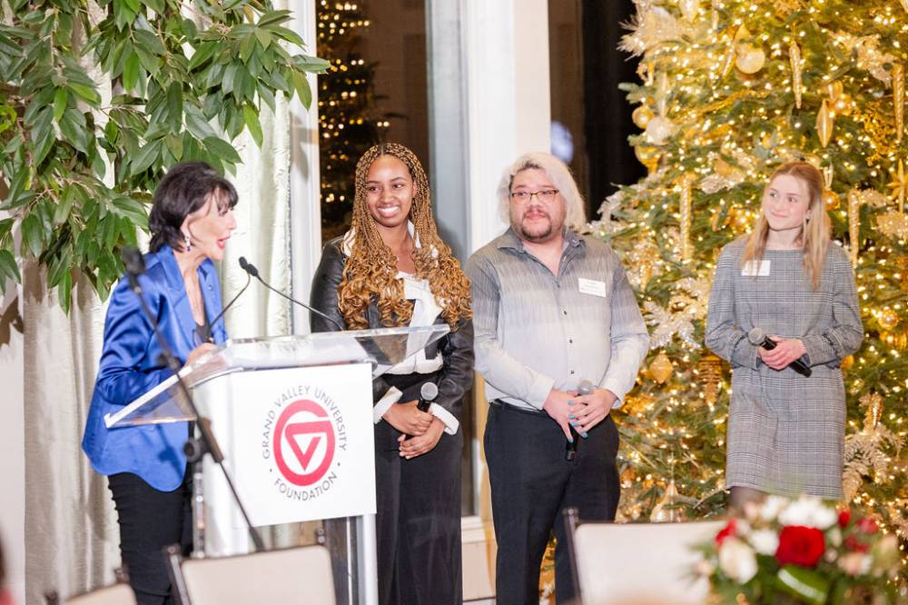 President Mantella speaking at podium with three GV students standing to her right, looking at her during her speech